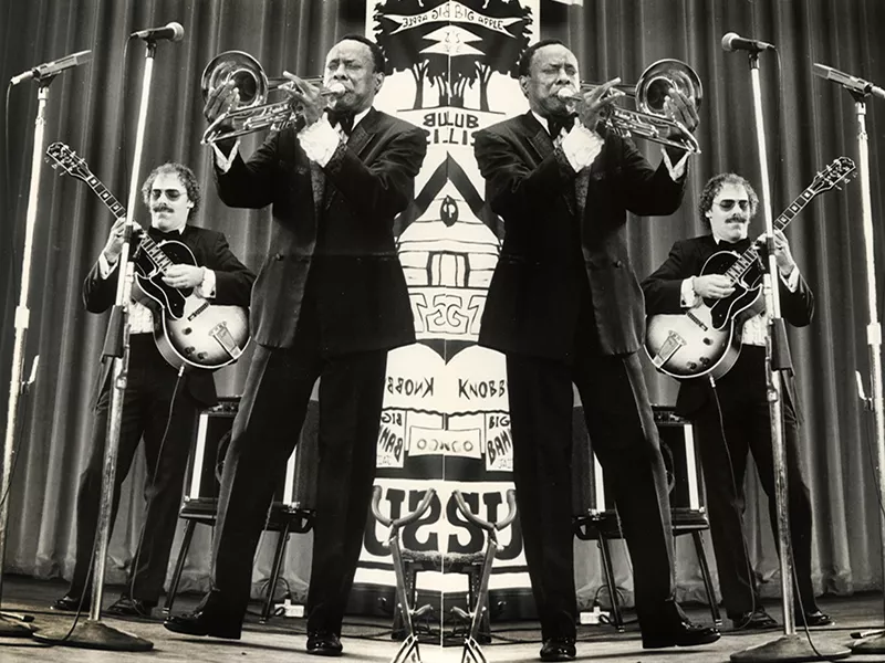 Mirrored image of two men performing, one is playing the guitar and the other is playing the trombone.