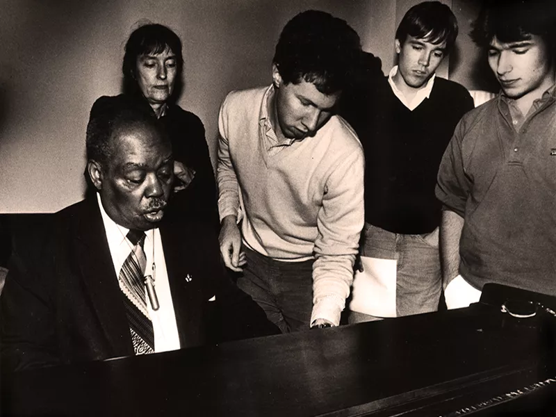 Sammy Price playing the piano with four others gathered around him.
