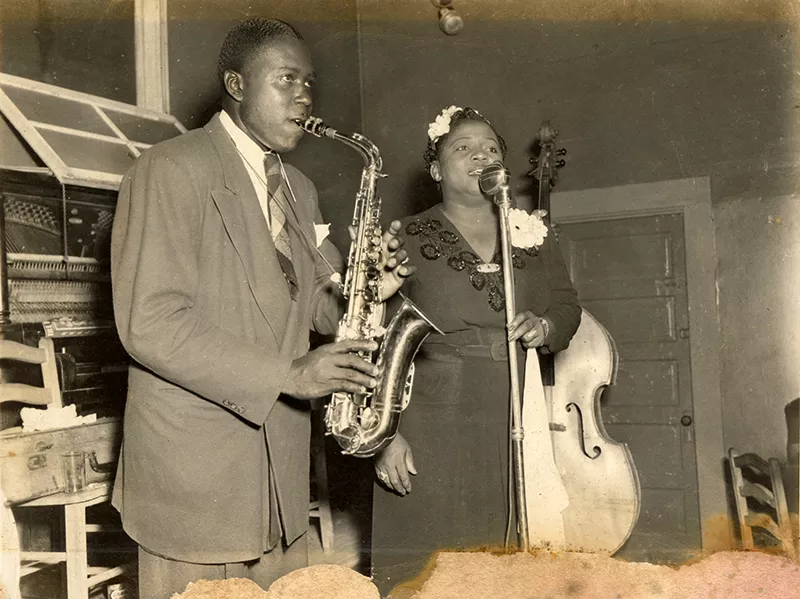 I.H. Smalley playing the saxophone next to a woman singing at a microphone.