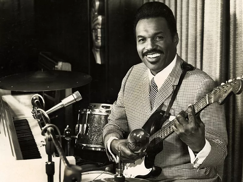 Lester Williams sitting in front of a piano with a guitar in his lap.